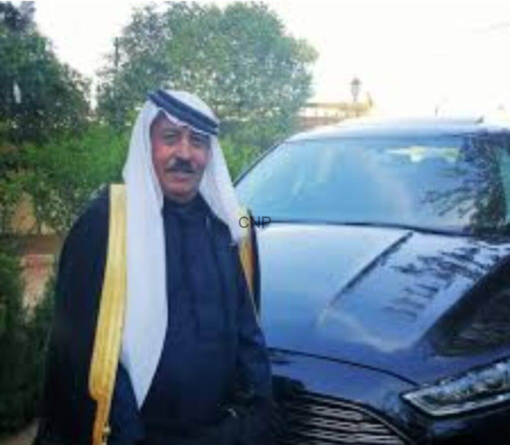 Man in traditional Middle Eastern attire (white headscarf and dark robe) standing beside a black SUV outdoors.