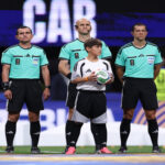 Line of soccer referees in teal shirts standing on the field with a child holding a ball at center, pre-match moment