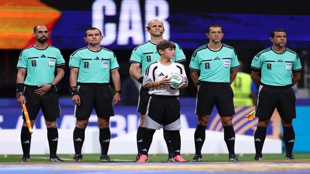 Line of soccer referees in teal shirts standing on the field with a child holding a ball at center, pre-match moment
