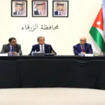 Panel of officials seated at a long black-draped table in a formal meeting, Jordanian flags on both sides and microphones in front of each person.