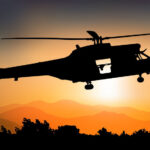Silhouette of a helicopter flying at sunset over a ridge and trees at dusk.