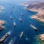 Aerial view of a busy shipping canal with several cargo ships moving through turquoise water between steep brown cliffs.