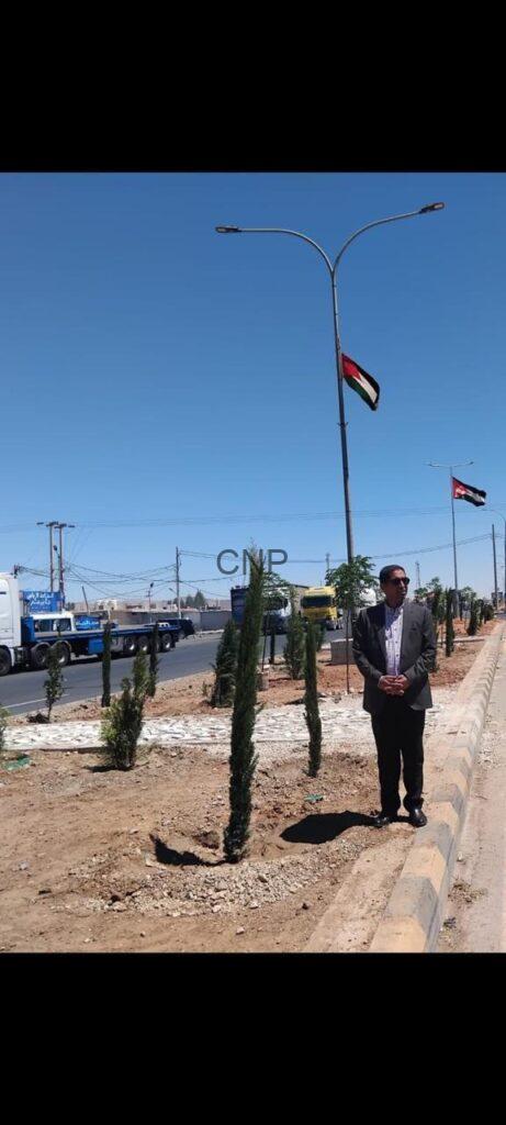 Man in a suit stands by a landscaped street median with small trees; a flag on a lamppost and trucks are visible in the background on a sunny day.