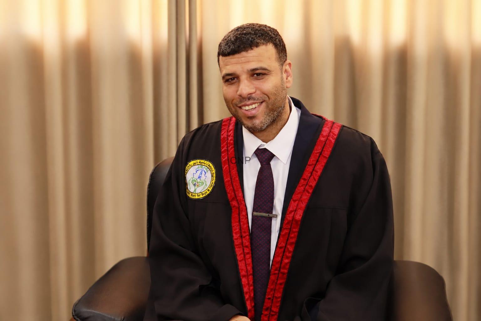 Smiling man in a black graduation gown with red trim and a white shirt, seated indoors against a light curtain backdrop.