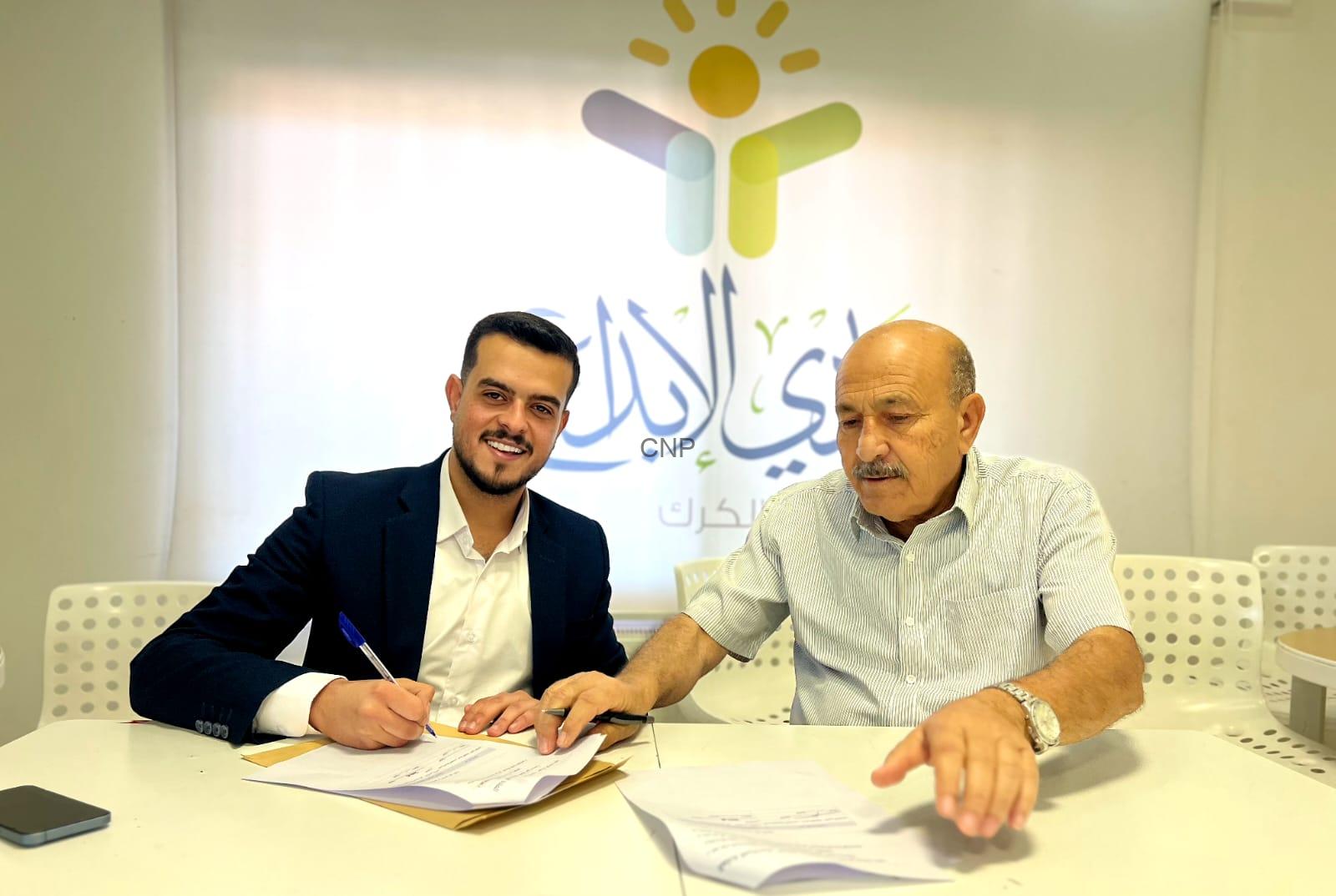 Two men sit at a white table signing documents, one in a suit and the other in a striped shirt, with a colorful logo on the wall behind them.