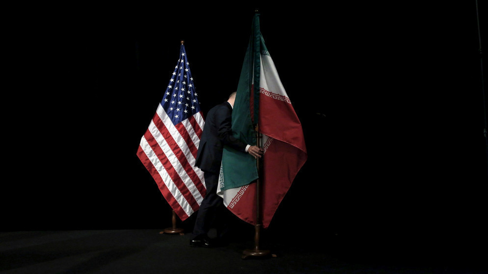 image - Center News Network Two flagpoles with the United States and Iran flags on a dark stage, a person in a suit between them holding the Iranian flag.