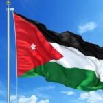 Jordanian flag fluttering on a flagpole against a bright blue sky, featuring a red triangle with a white star.