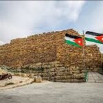 Stone fortress wall with the letters 'CNF' carved into it, flanked by two Jordanian flags on a doorway under a cloudy sky.