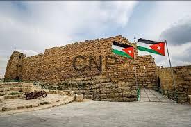 Stone fortress wall with the letters 'CNF' carved into it, flanked by two Jordanian flags on a doorway under a cloudy sky.