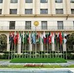 Front of a government building with a gold emblem above the entrance, a row of national flags on flagpoles, and a reflecting pool in the foreground.