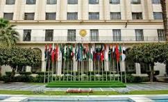 Front of a government building with a gold emblem above the entrance, a row of national flags on flagpoles, and a reflecting pool in the foreground.
