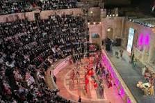 Indoor arena with a circular performance pit and tiered audience seating, performers in red on a brightly lit stage surrounded by spectators.