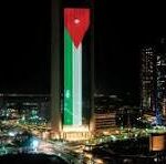 Nighttime city skyline with a tall, illuminated green-and-white tower topped with a red emblem, surrounded by other buildings.