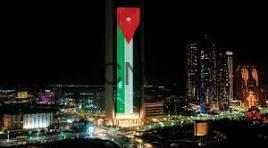 jordan - Center News Network Nighttime city skyline with a tall, illuminated green-and-white tower topped with a red emblem, surrounded by other buildings.