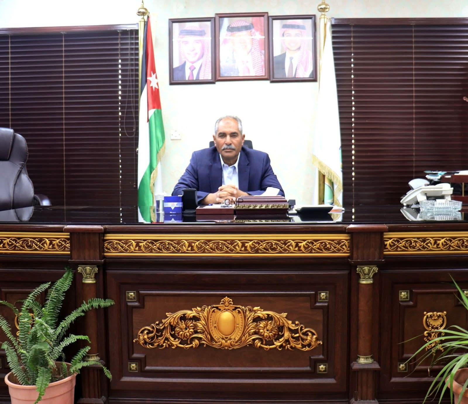 khal - Center News Network Man in a blue suit seated at a large ornate wooden desk in a formal office, with a Jordan flag and framed portraits on the wall behind him.