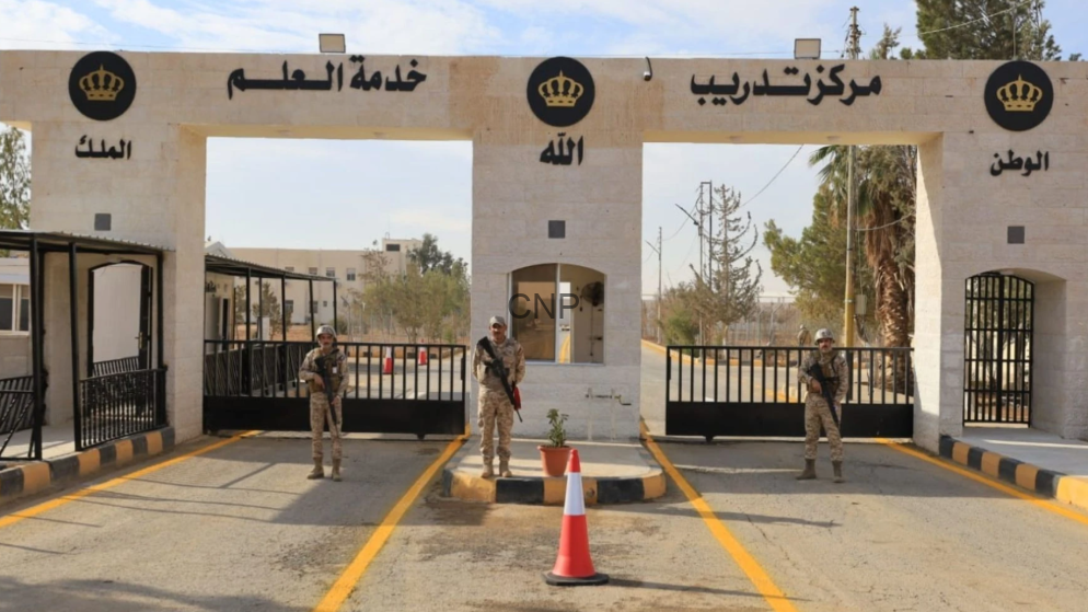 Border checkpoint entrance with three arches, armed soldiers standing guard, a small guardhouse between arches, yellow lane markings, and an orange traffic cone; Arabic signs and crown logos on the facade.