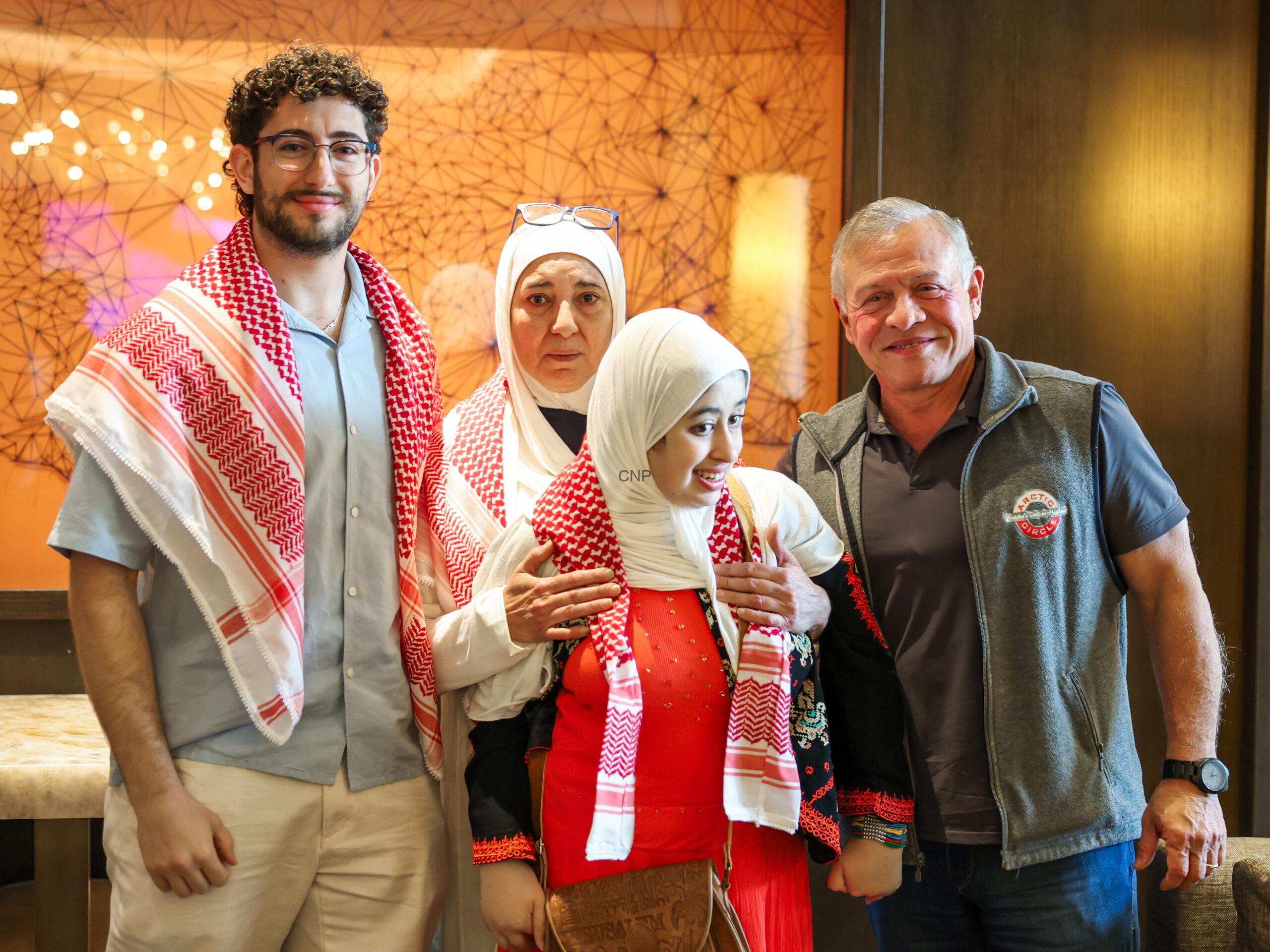 Four people posing together indoors; two men on the ends wear casual clothes and red-and-white keffiyehs, while two women in white hijabs stand between them against an orange patterned wall.