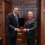 Two men in suits exchange a red velvet medal box, smiling, in a wood-paneled room.