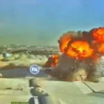 Aerial view of a large explosion on a rooftop facility, with bright orange flames and thick black smoke rising from a crater, under a clear blue sky.