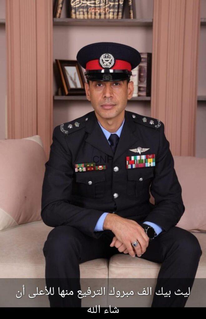 Portrait of a male military officer in full dress uniform with medals, seated on a beige sofa, wearing a peaked cap with a red band, in a room with shelves behind him; hands folded in his lap and a badge on the chest are visible.