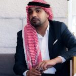 Man seated on a sofa wearing a red-and-white keffiyeh and a dark blazer, looking toward the camera in a formal pose.