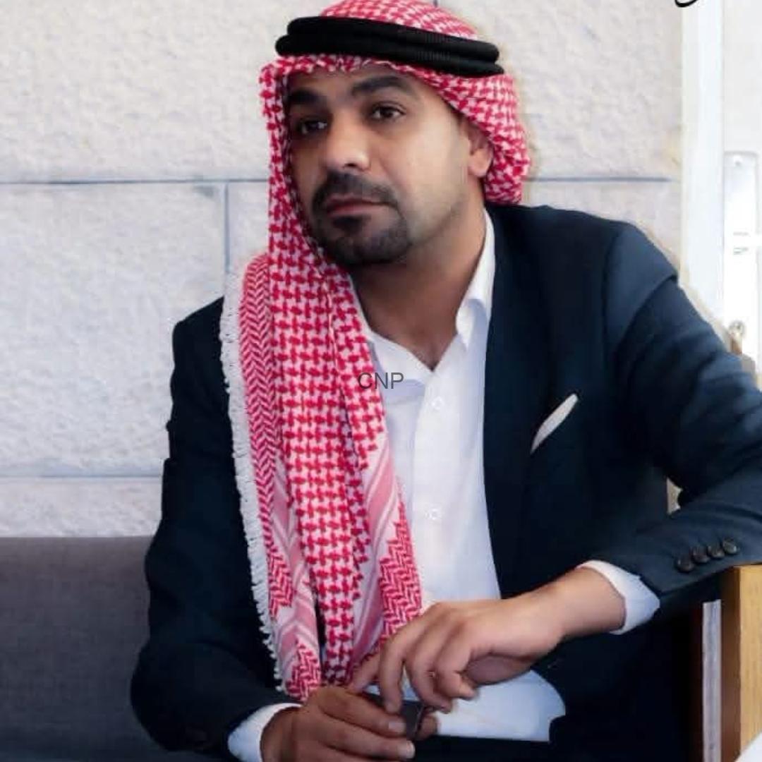 Man seated on a sofa wearing a red-and-white keffiyeh and a dark blazer, looking toward the camera in a formal pose.