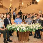 Group of officials and military officers seated at a formal ceremony around a flower-adorned table in a grand hall.