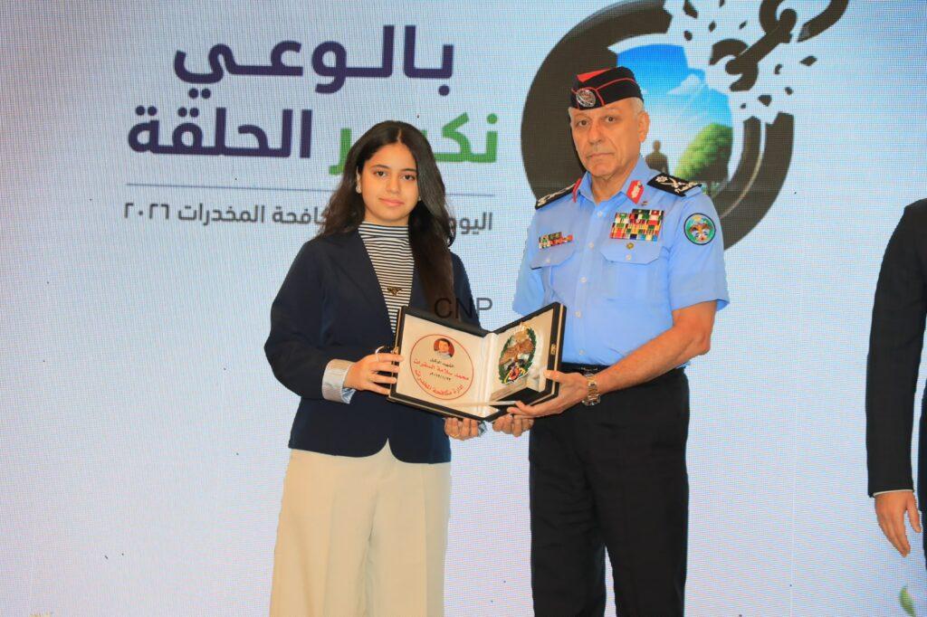 mok2 - Center News Network Young woman in a navy blazer receives a certificate from a uniformed officer at an awards ceremony on stage, with Arabic text backdrop.