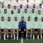 Group photo of a soccer team posing on a stadium field in matching light green training jackets; a coach in a dark tracksuit sits center front, bleachers and a 'Portland' sign are in the background.