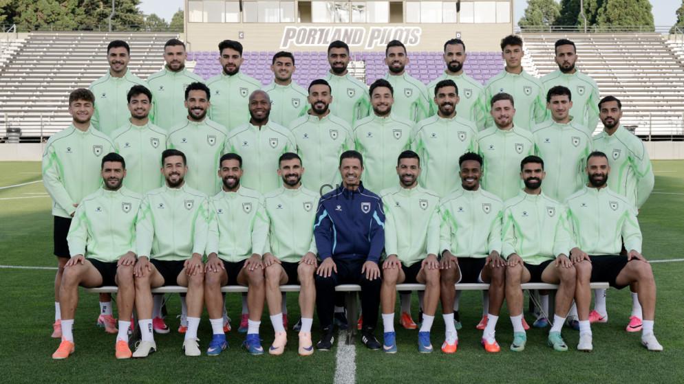 Group photo of a soccer team posing on a stadium field in matching light green training jackets; a coach in a dark tracksuit sits center front, bleachers and a 'Portland' sign are in the background.