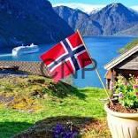 Norwegian flag by a lakeside cabin, with flowers, a boat on the fjord and snow-capped mountains in the background.
