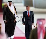 Two men walk down a red carpet together, one in traditional white-and-black robes with a head covering and the other in a dark suit and red tie, as onlookers photograph nearby.