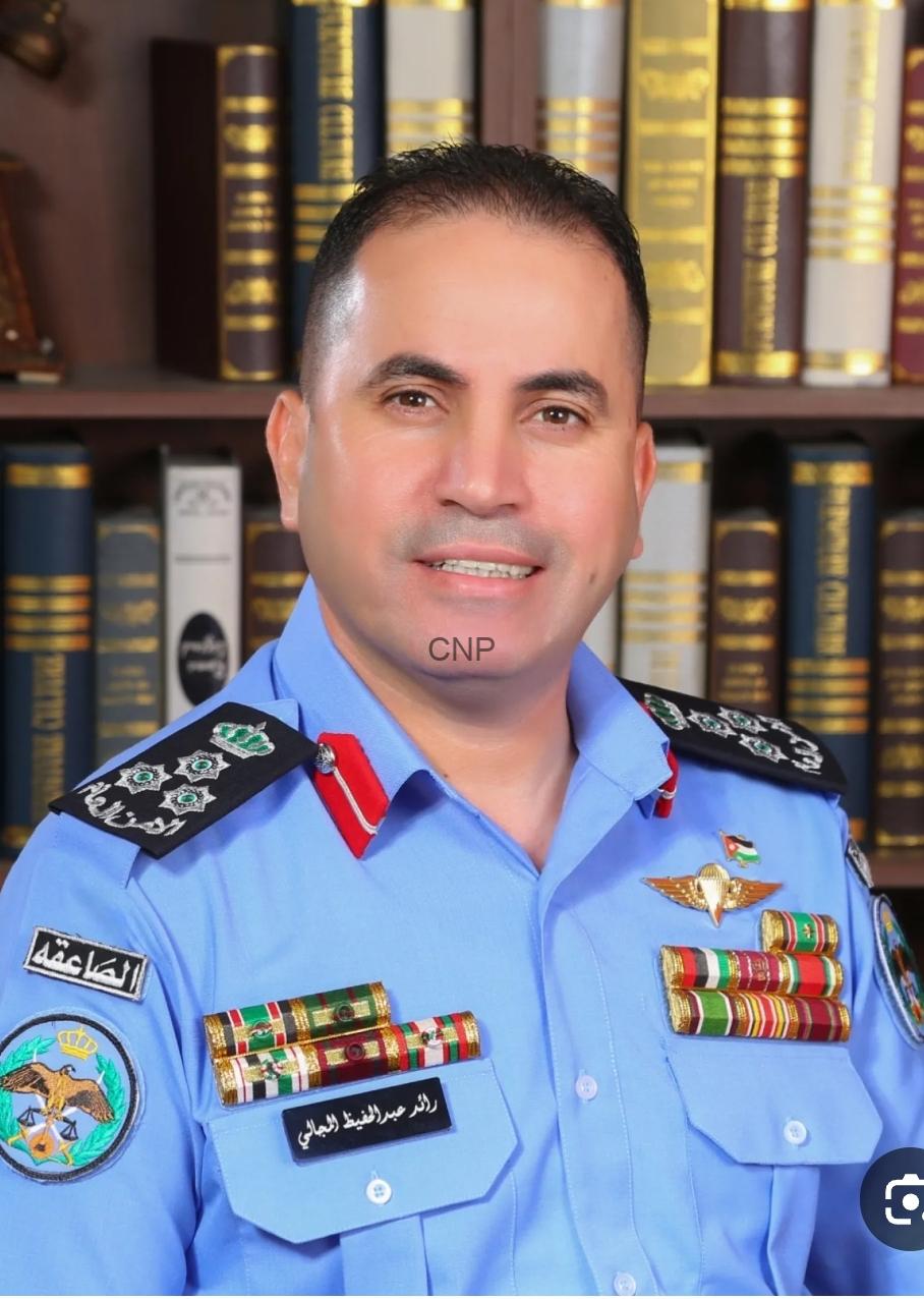 Portrait of a male officer in a light blue uniform with multiple ribbons and insignia, standing in front of a bookshelf full of legal tomes for an official portrait
