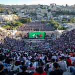 Thousands of spectators fill a sunlit outdoor amphitheater, watching a large televised football match on a central screen.