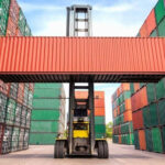 Forklift lifting a red shipping container between stacked multicolored containers in a cargo yard.