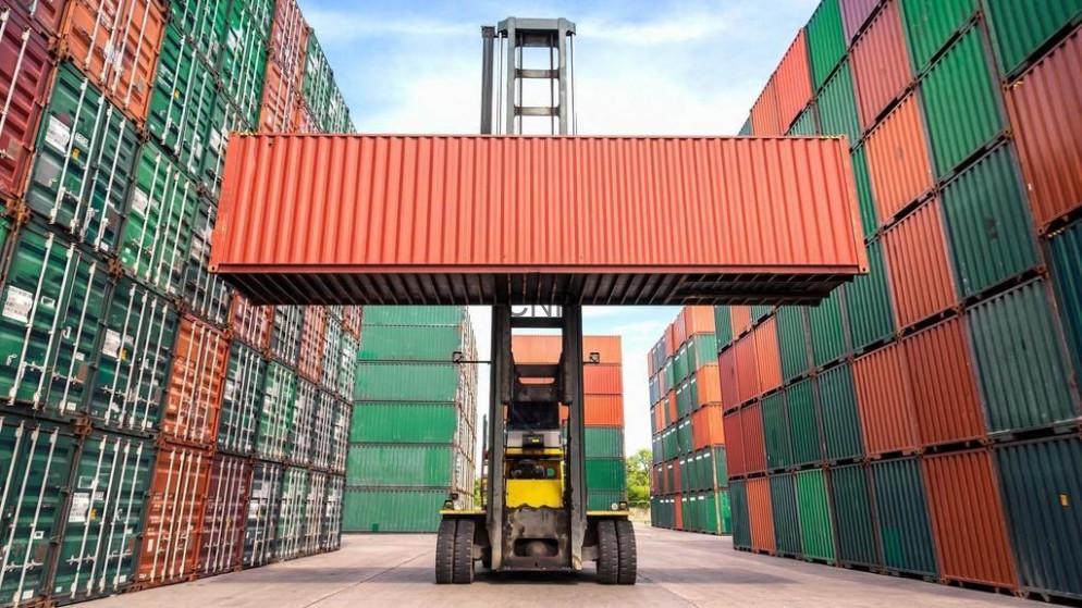 Forklift lifting a red shipping container between stacked multicolored containers in a cargo yard.