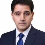 Professional head-and-shoulders portrait of a man in a navy suit and blue tie with neutral expression, white background.