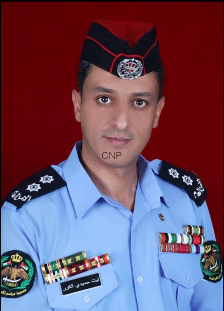 Portrait of a man in a light blue ceremonial uniform with medals and shoulder insignia, wearing a black cap with red trim against a red background.