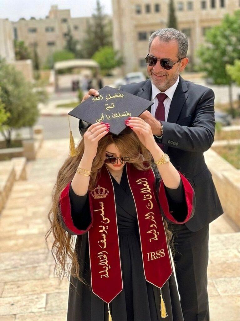 Man in a suit and sunglasses places a graduation cap on a woman in a black dress and maroon stole outdoors on a campus-like walkway, cap with gold Arabic text visible.