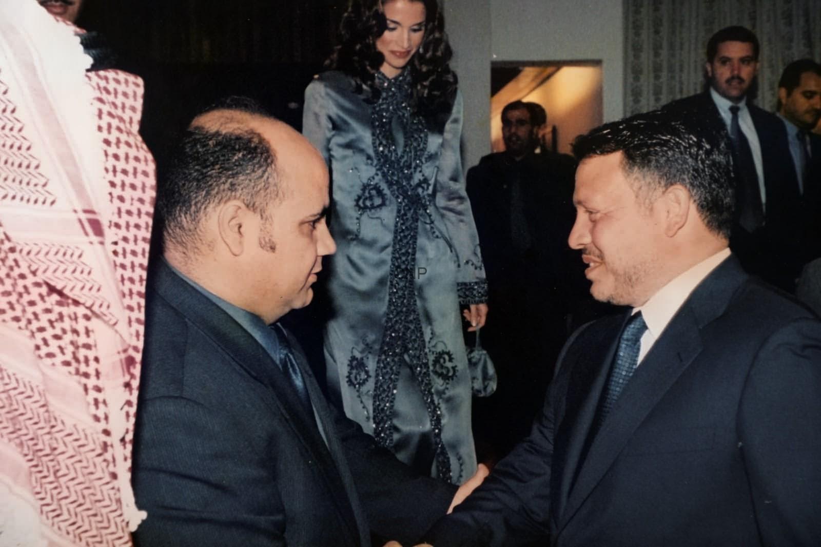 Two men in dark suits shake hands at a formal event, with a woman in a blue embellished outfit visible in the background.