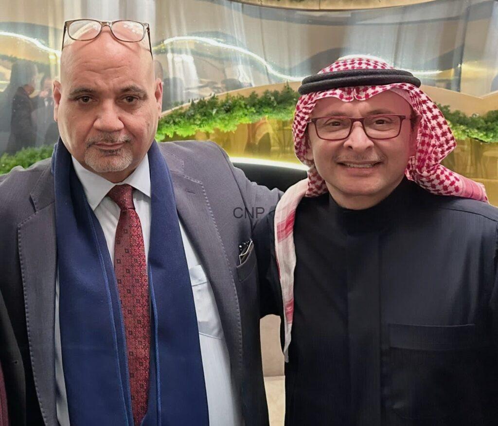 Two men posing for a photo indoors; left man in suit and red tie with glasses on his head, right man in black thobe and red‑white keffiyeh, both smiling.