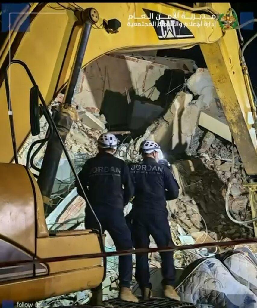 Two rescue workers in dark uniforms and white helmets stand amid rubble from a collapsed building, next to a yellow excavator arm during a nighttime search.