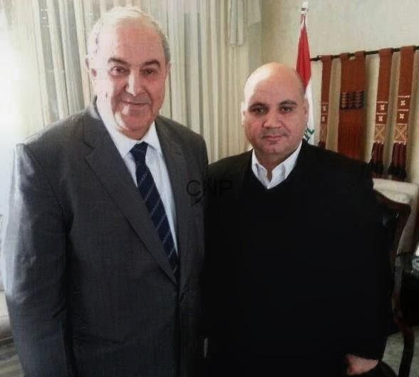 Two men in suits pose for a formal photo indoors, with a Lebanese flag visible in the background.