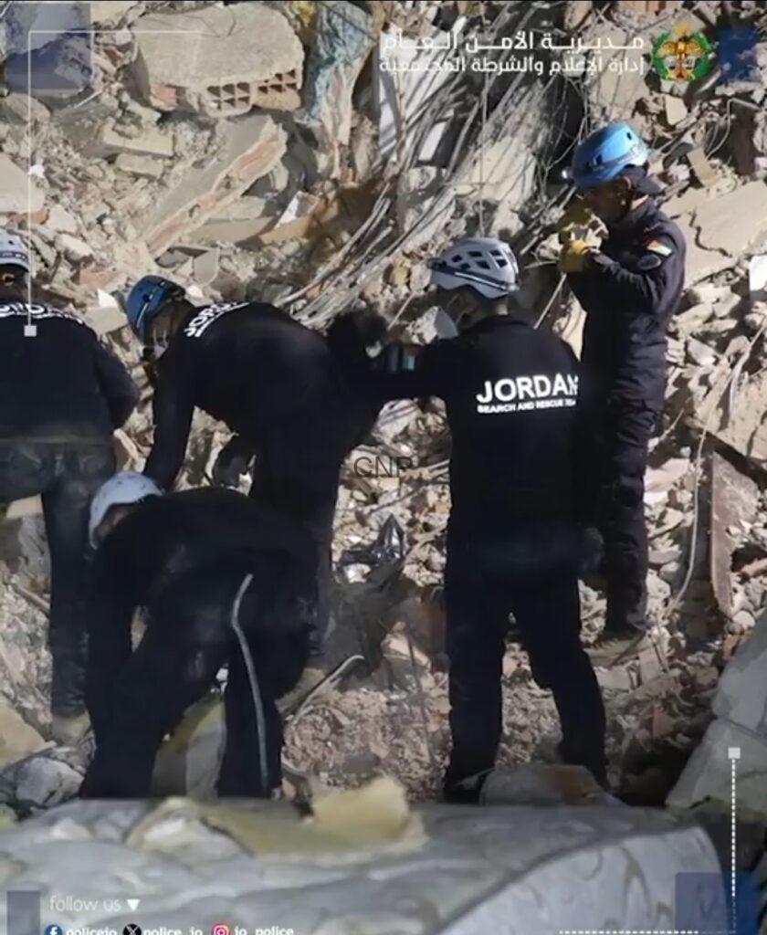 Rescue workers in black uniforms and helmets search debris in a collapsed building, one labeled Jordan Search and Rescue, amid tangled rebar and rubble