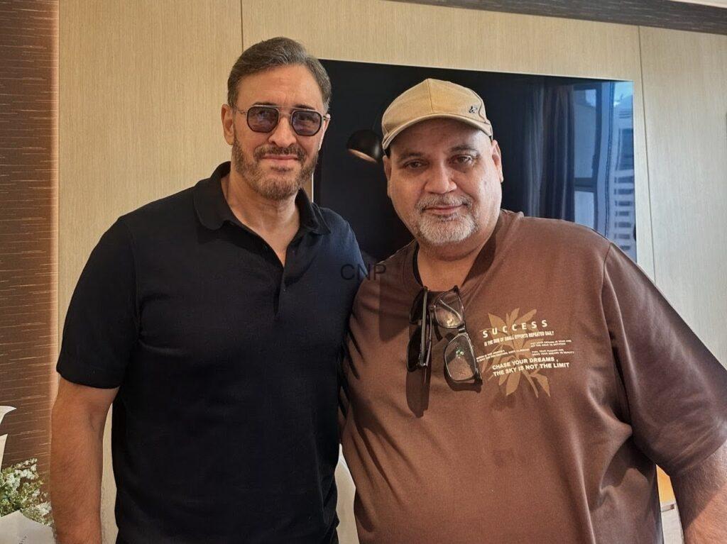 Two men pose indoors for a photo; one in a black polo with sunglasses, the other in a brown sweatshirt and tan cap, both smiling.