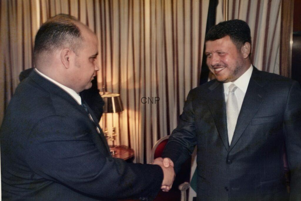 Two men in dark suits shaking hands in an indoor setting, smiling as they greet each other.