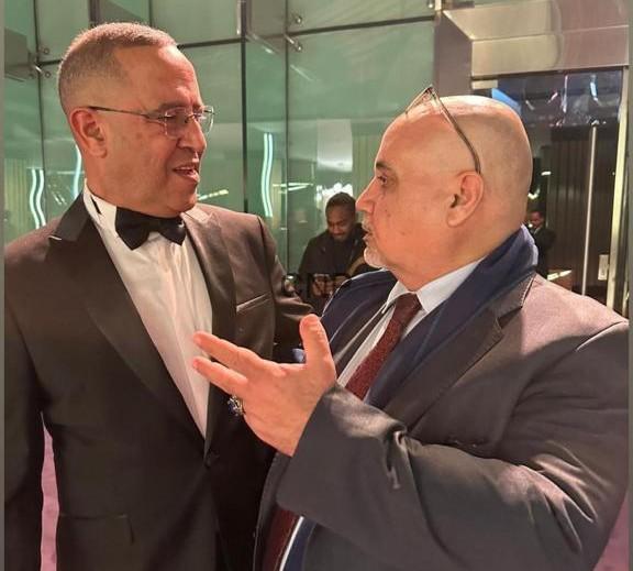 Two men in formal attire converse in a glass-walled lobby; one gestures with his hand.