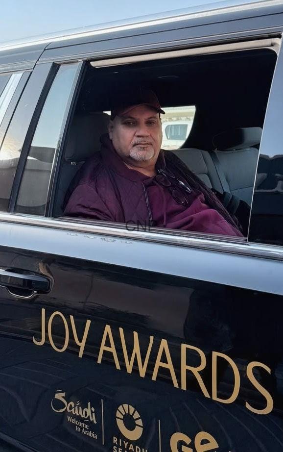 Man in a maroon jacket sits in a black car window beside 'JOY AWARDS' on the vehicle's exterior.