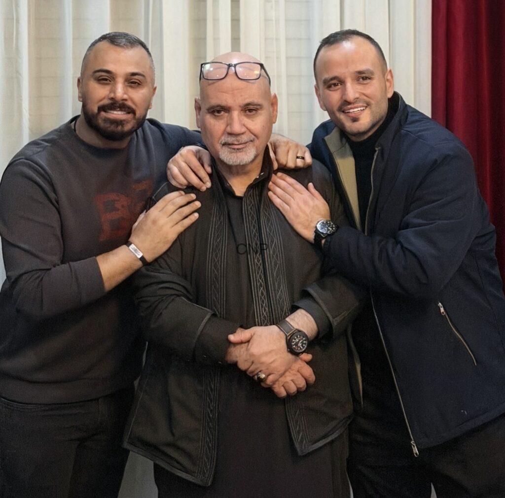 Three men posing closely for a group photo, with the middle older man wearing a black traditional outfit and glasses on his head, flanked by two smiling friends with their arms around him.