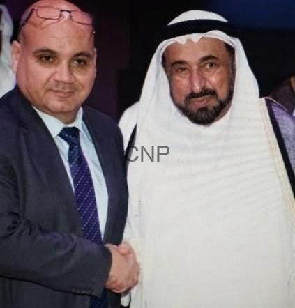 Two men in formal attire shaking hands: one in a suit and tie, the other in a white traditional thawb and head covering, both smiling slightly.